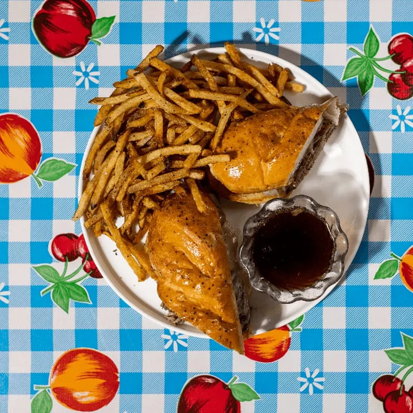 A Doo-Dah Diner French Dip sandwich with fries and au jus on a blue and white checkered tablecloth.